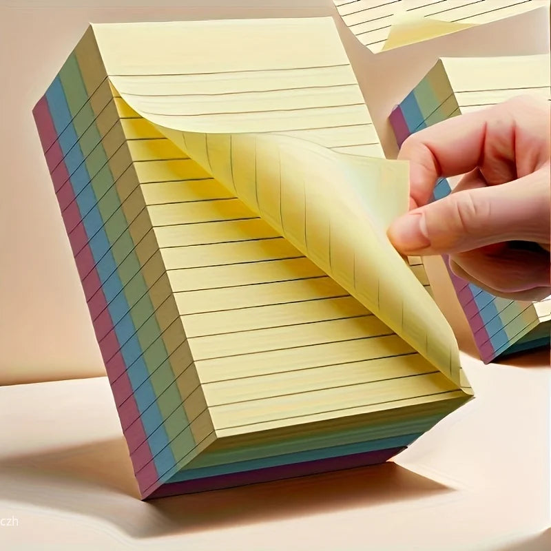 Box of lined sticky note pads with layered pastel sheets; hand holding an edge.